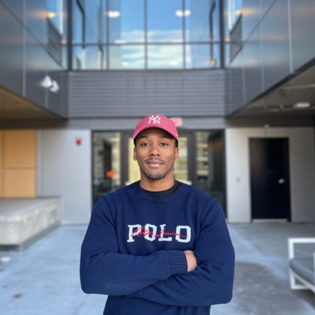 Cannon, wearing a coral-red baseball cap and a navy knit sweater, standing outdoors with arms crossed.