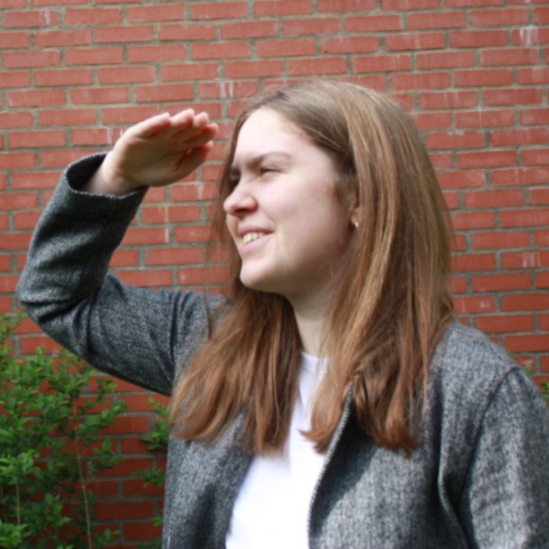 Daria, long light-brown hair, wearing a grey tweed jacket over a white shirt, photographed in profile against a brick wall.